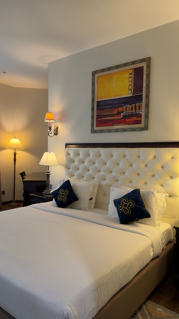 gallery-04 Luxurious hotel room featuring a plush bed, decorative pillows, and modern lighting for a cozy stay.